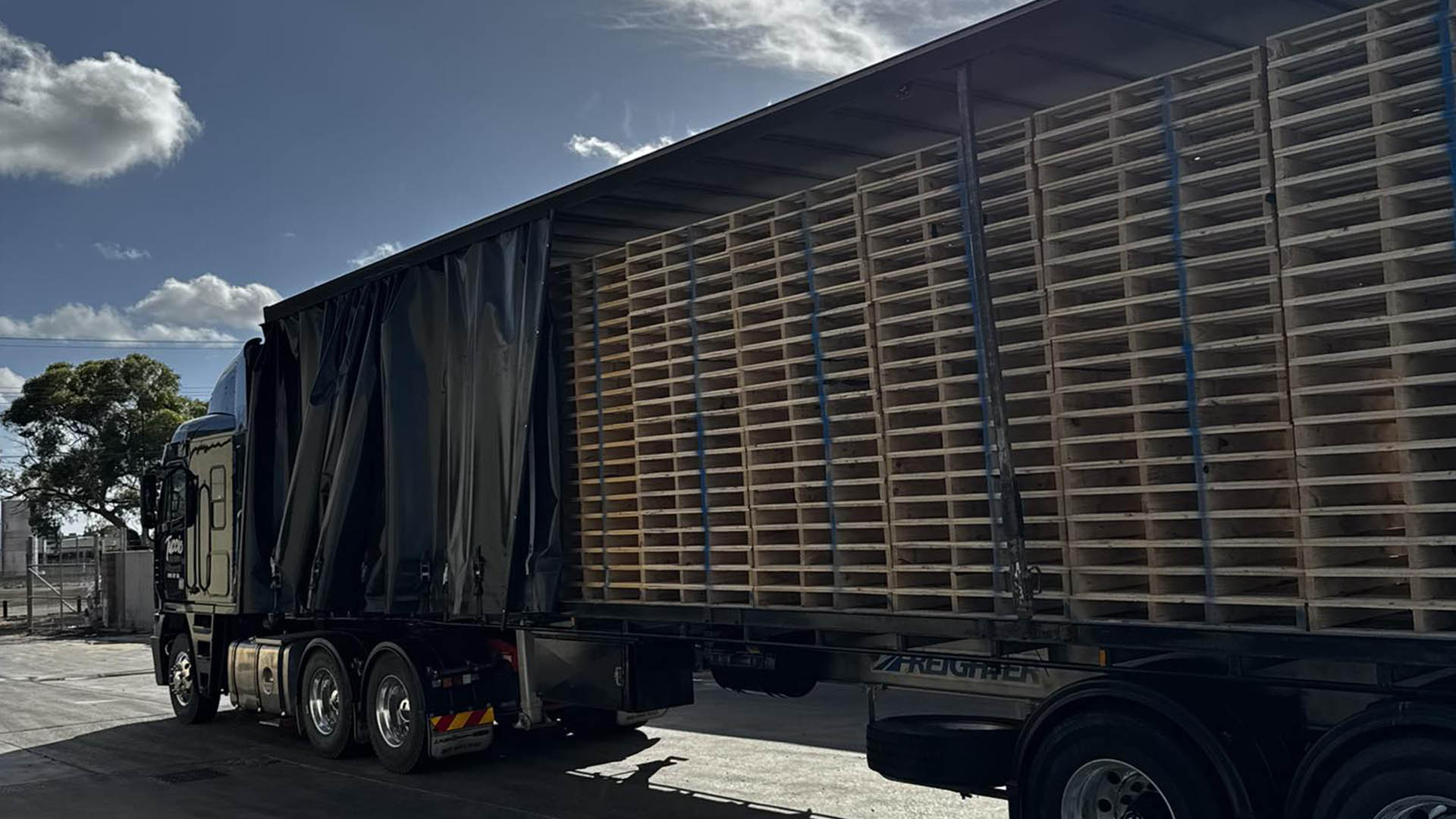 Wide view of a curtain-sider semi-trailer with curtains pulled back, loaded with timber pallets